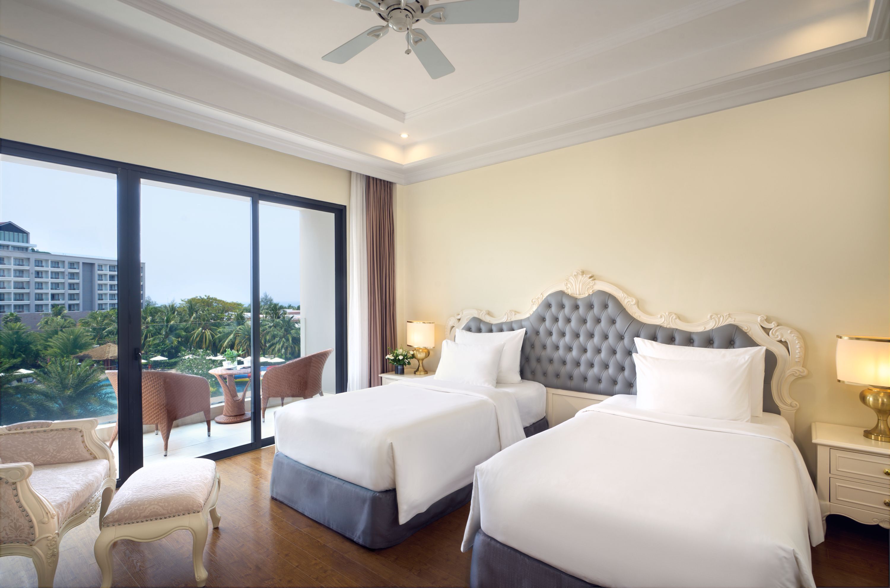 Ein Hotelzimmer mit zwei Betten, einem großen Fenster mit Blick auf ein Gebäude und Palmen, einem Deckenventilator und einem kleinen Tisch mit Lampe.