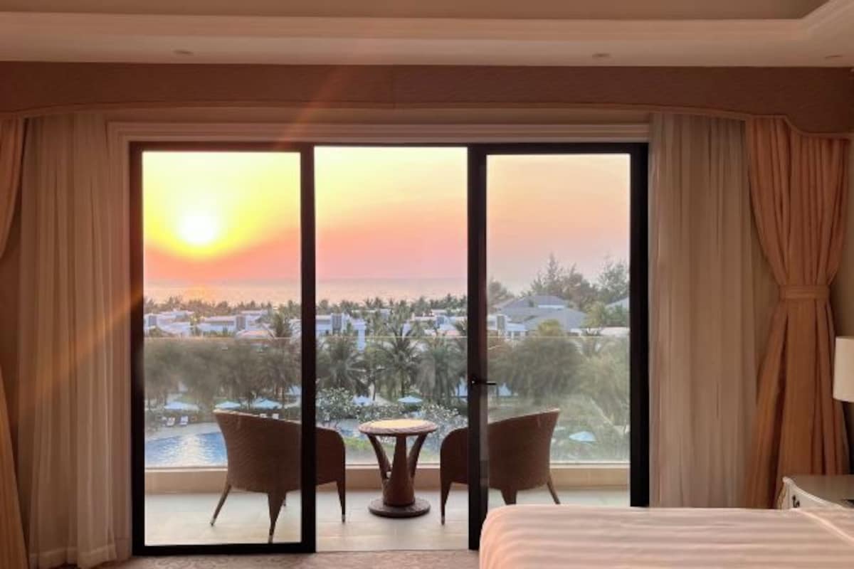 Habitación de hotel con un amplio ventanal corredero que da a un balcón con vistas a un complejo turístico y al atardecer.