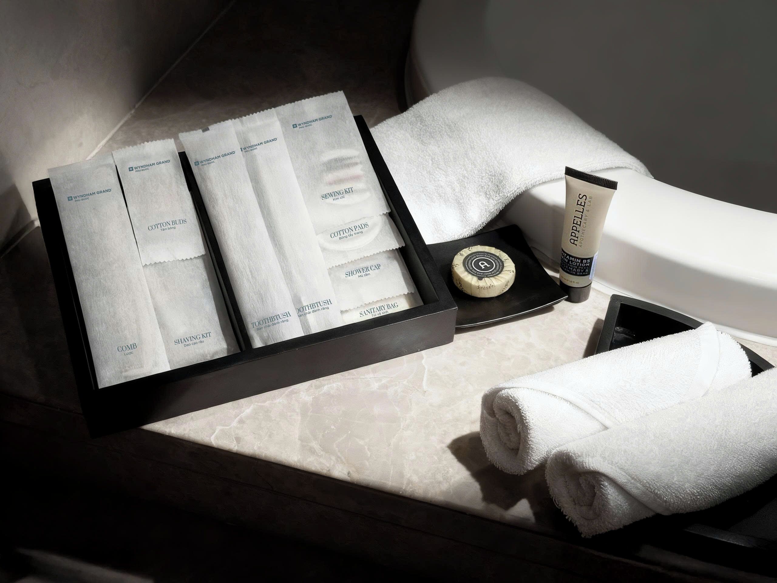 A neatly arranged bathroom with a black tray holding a shaving kit, cotton buds, a toothbrush, a comb, a sanitary bag, and a shower cap.