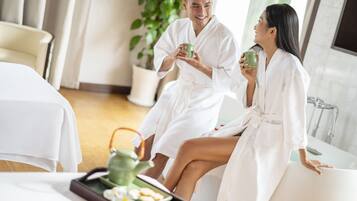 A couple in white bathrobes sitting by a bathtub in a well-lit room with a plant and a tray of tea and cookies.