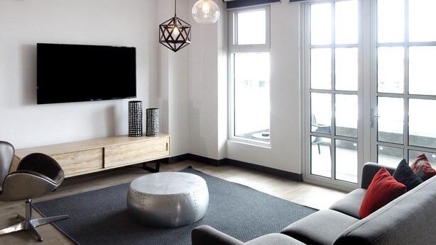A modern living room with a flat-screen TV, a wooden console, a round ottoman, and a grey sofa with red cushions.