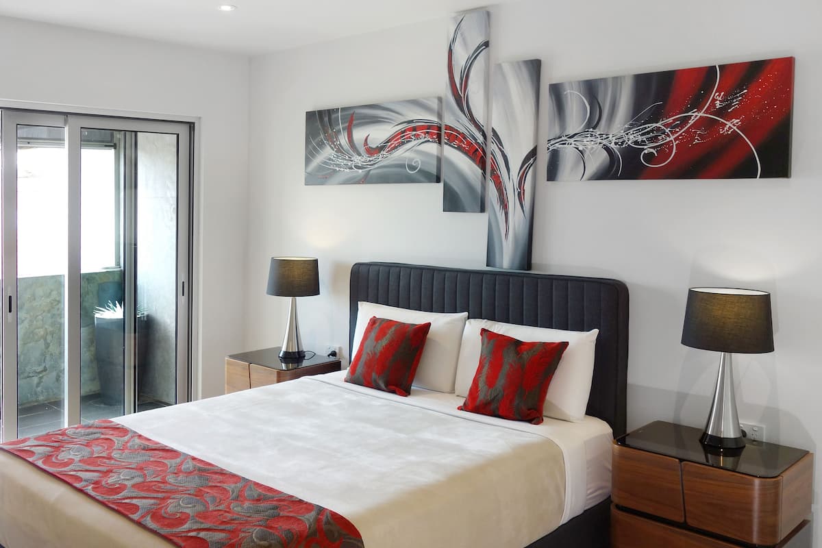 A modern hotel room with a large bed, two bedside tables, a nightstand, a balcony with a chair, and abstract wall art.