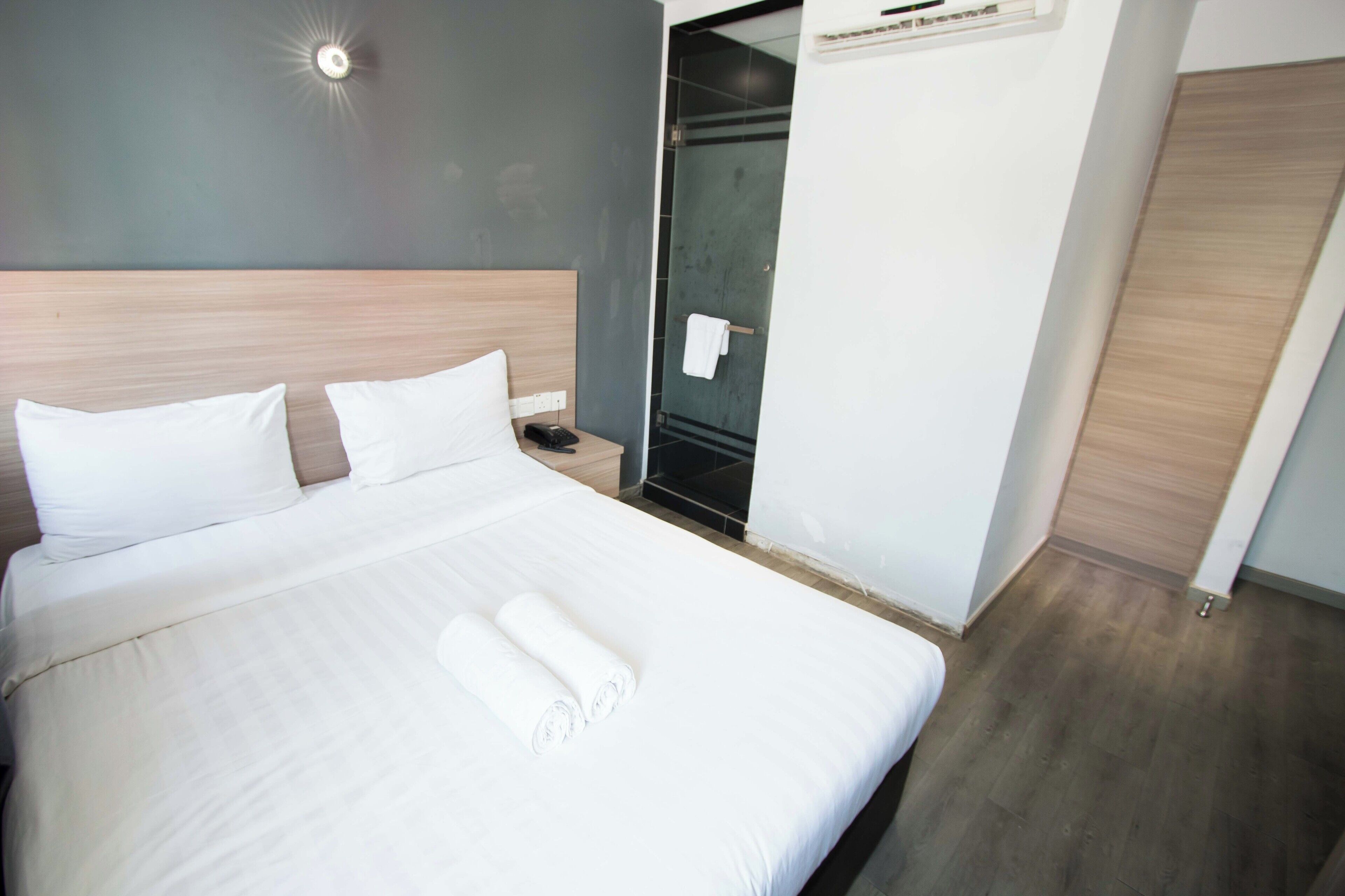 A neatly made bed with white linens and rolled towels, a bedside table with a phone, and a wall-mounted air conditioning unit.