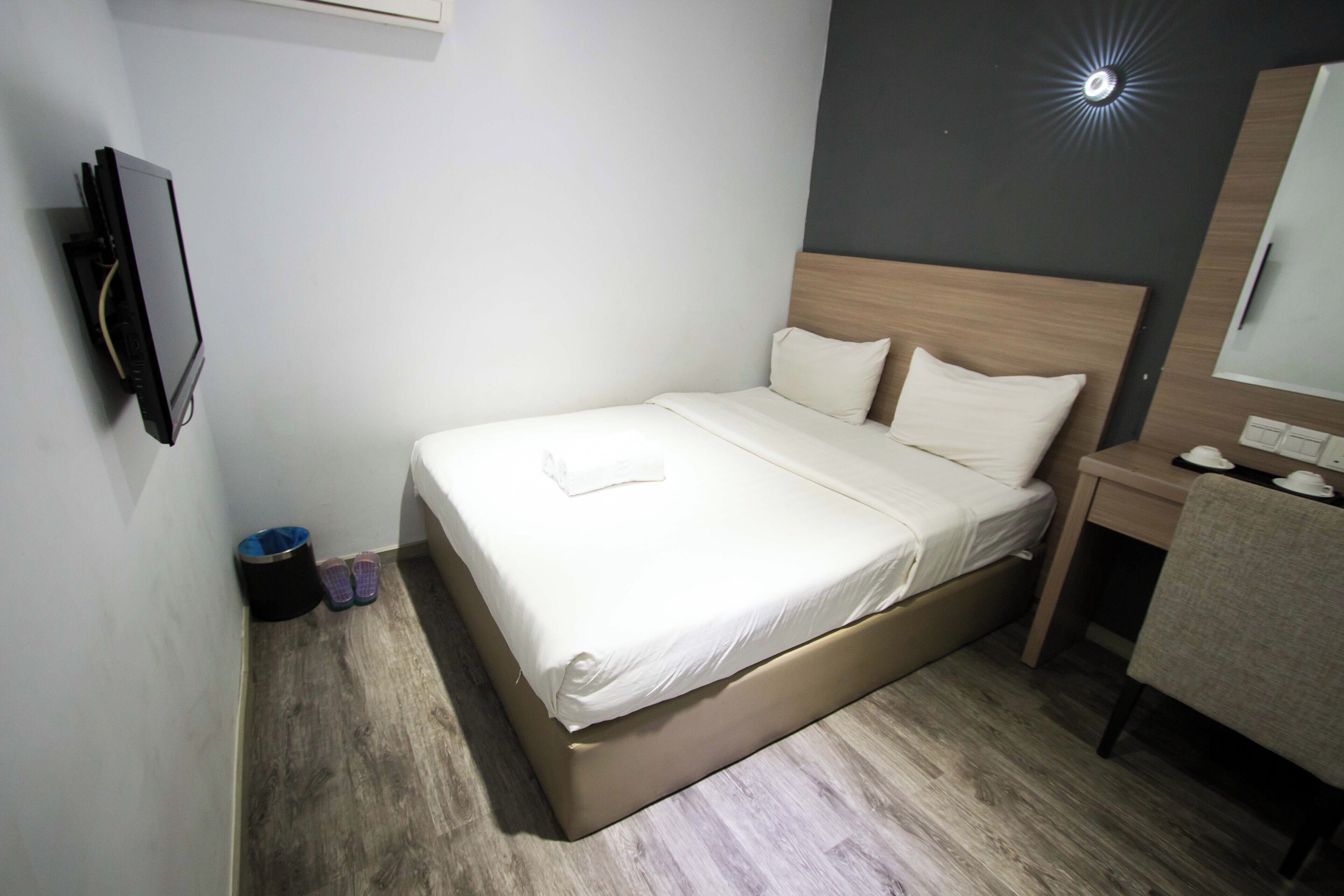 Una cama bien hecha con edredón blanco y almohadas, una mesita de noche con lámpara y un pequeño escritorio con silla.