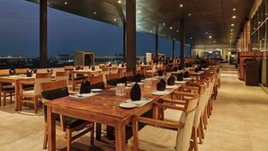 A rooftop restaurant with wooden tables and chairs, large windows, and a view of the city skyline at dusk. - Sayaji Raipur (Raipur)