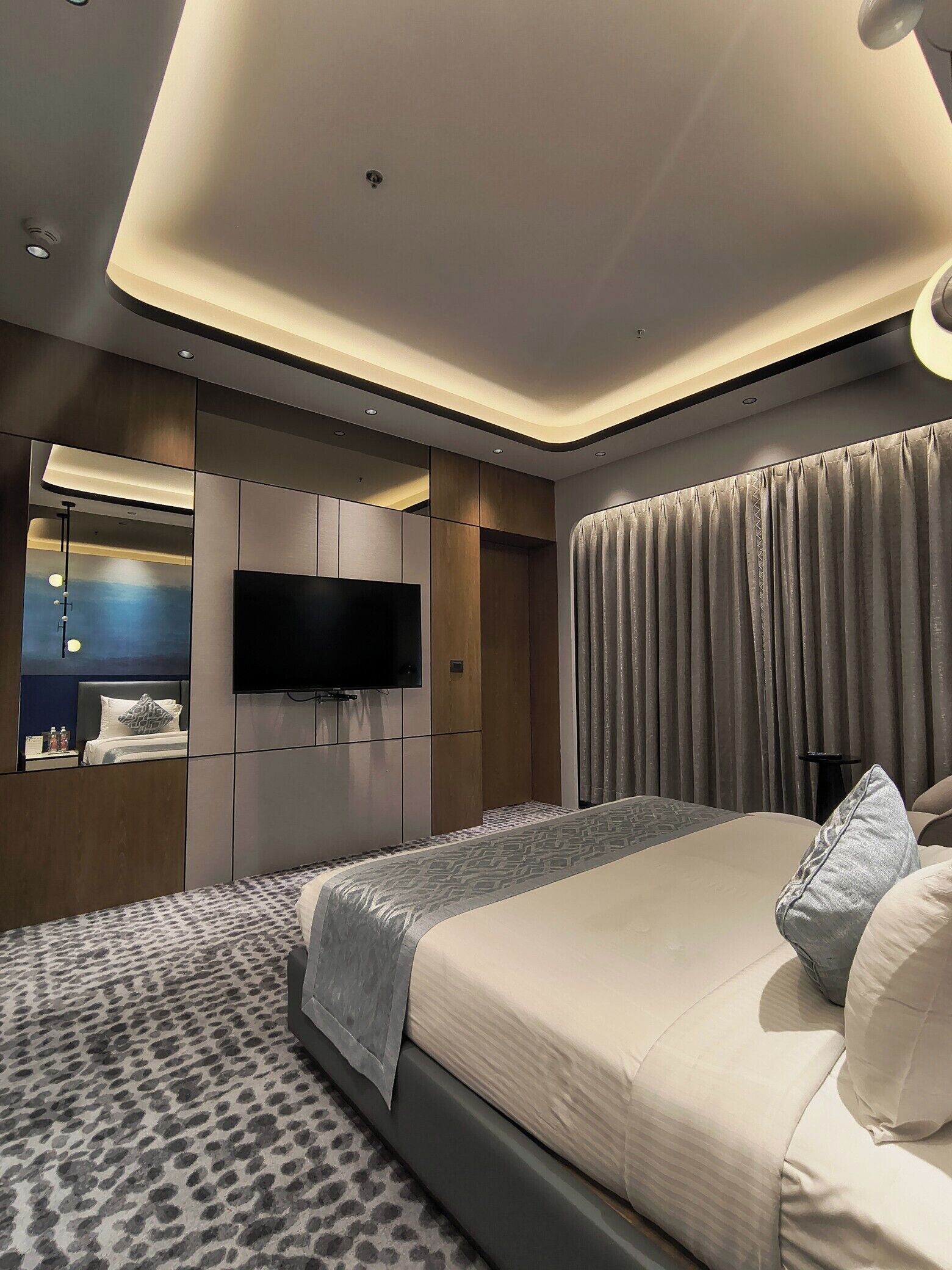 A modern bedroom with a large bed, a flat-screen TV mounted on the wall, and a ceiling with recessed lighting.