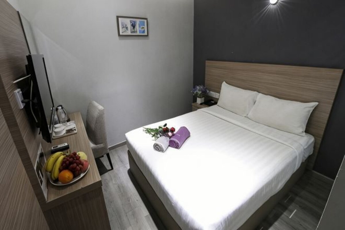 A hotel room with a bed, a bedside table with a fruit bowl, a TV, and a nightstand with a lamp.