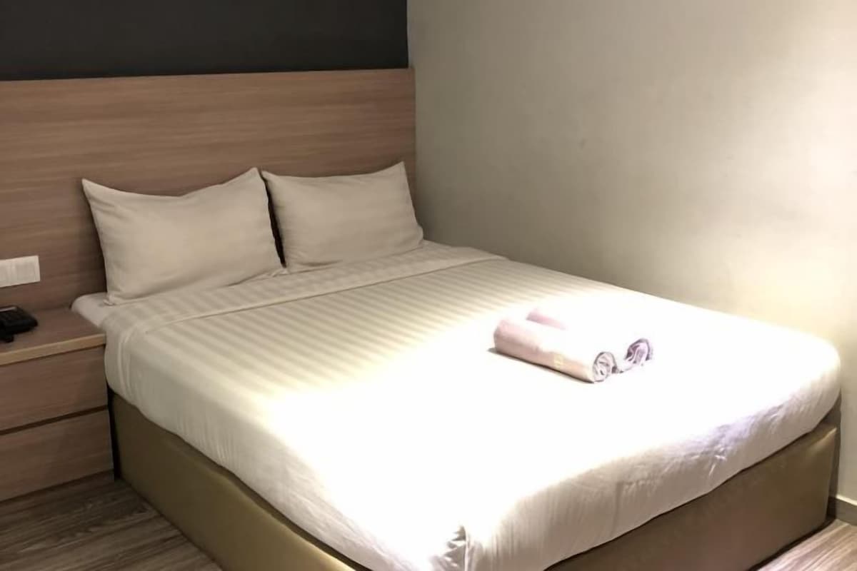 A neatly made bed with a white comforter and two pillows, a wooden headboard, and a bedside table with a phone and a small item on it.
