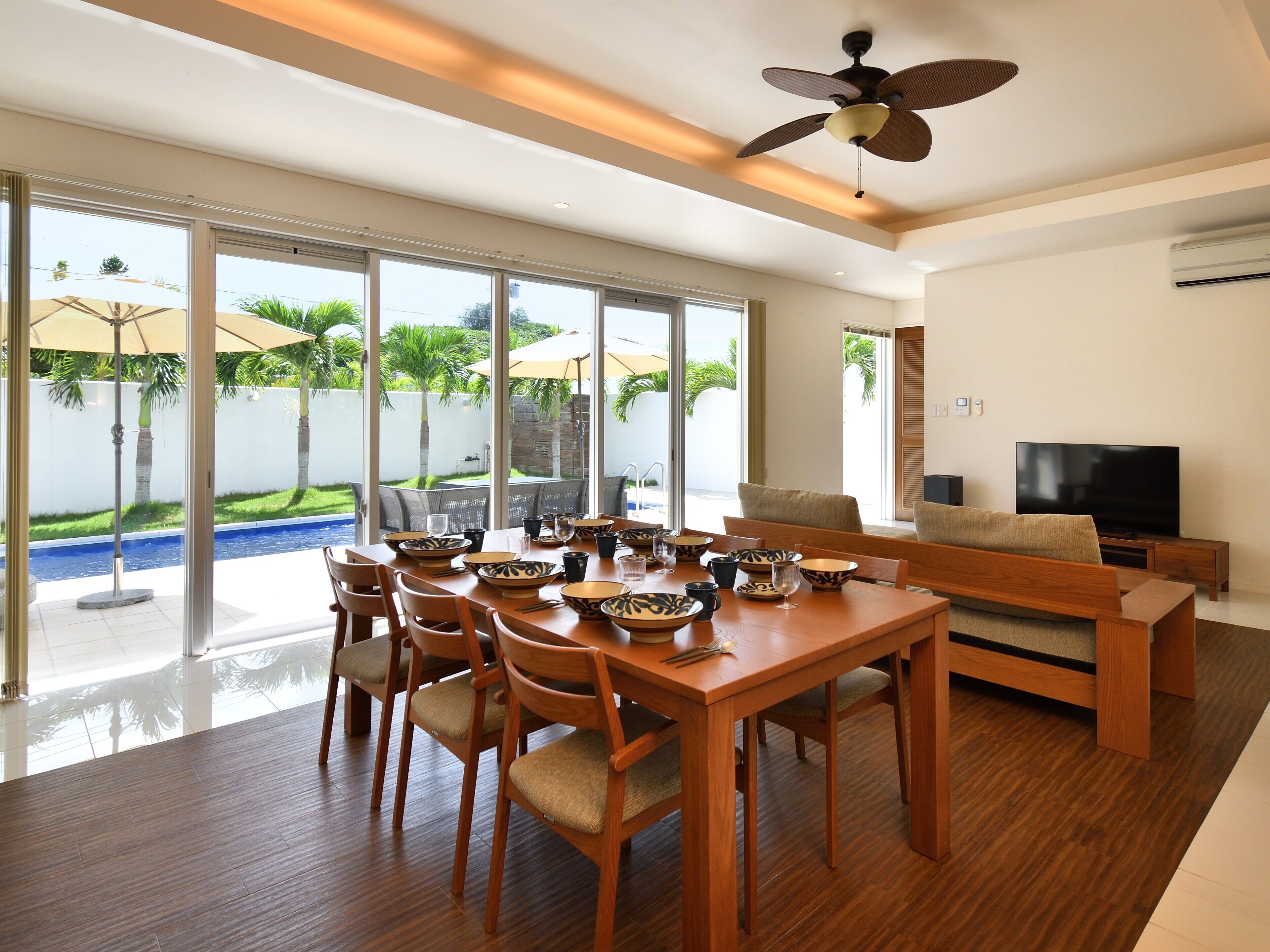 A spacious dining area with a wooden table and chairs, a ceiling fan, and a large window offering a view of a swimming pool and greenery.