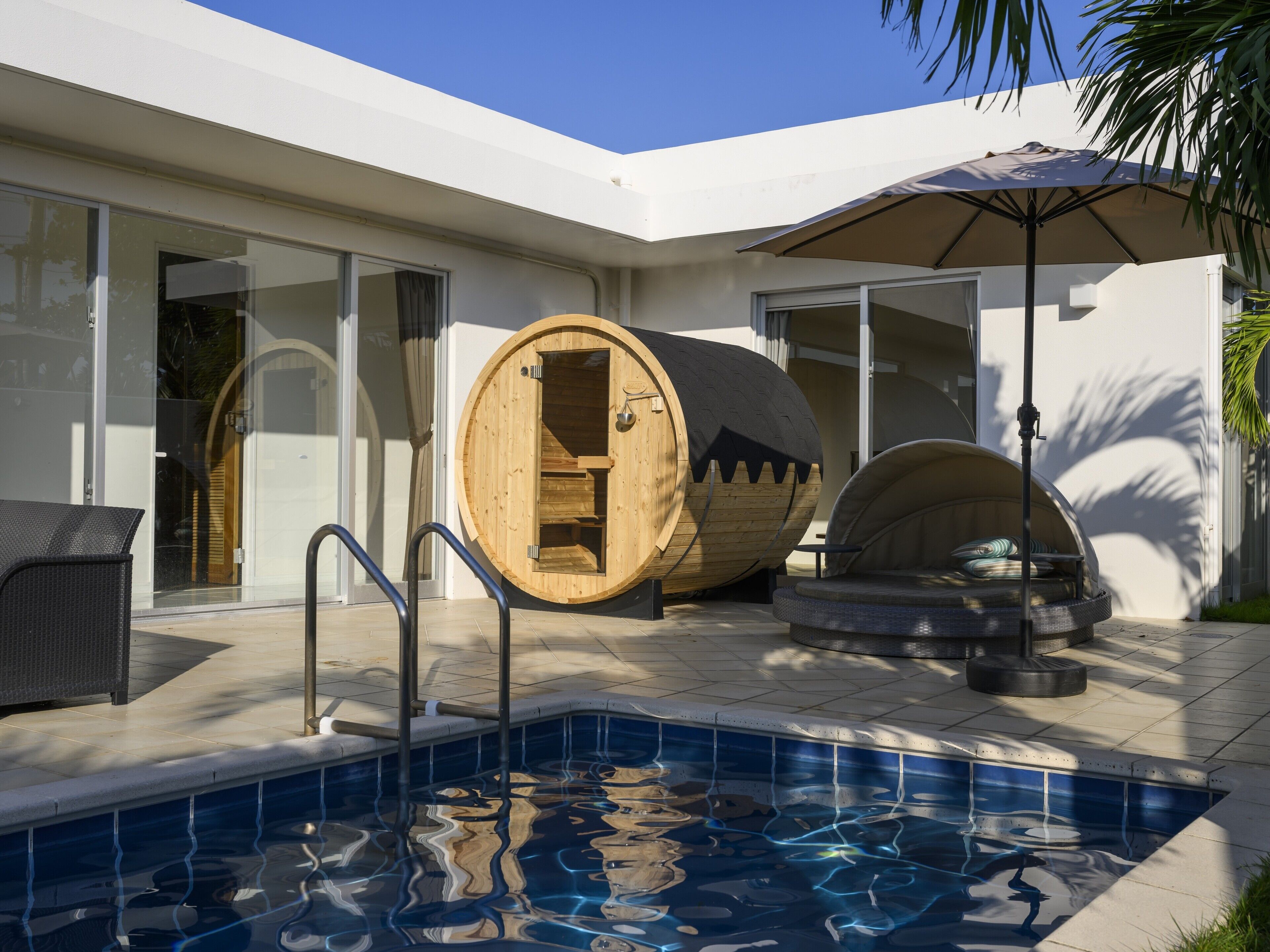A modern outdoor pool area with a wooden barrel sauna, a lounge chair, and a covered seating area.