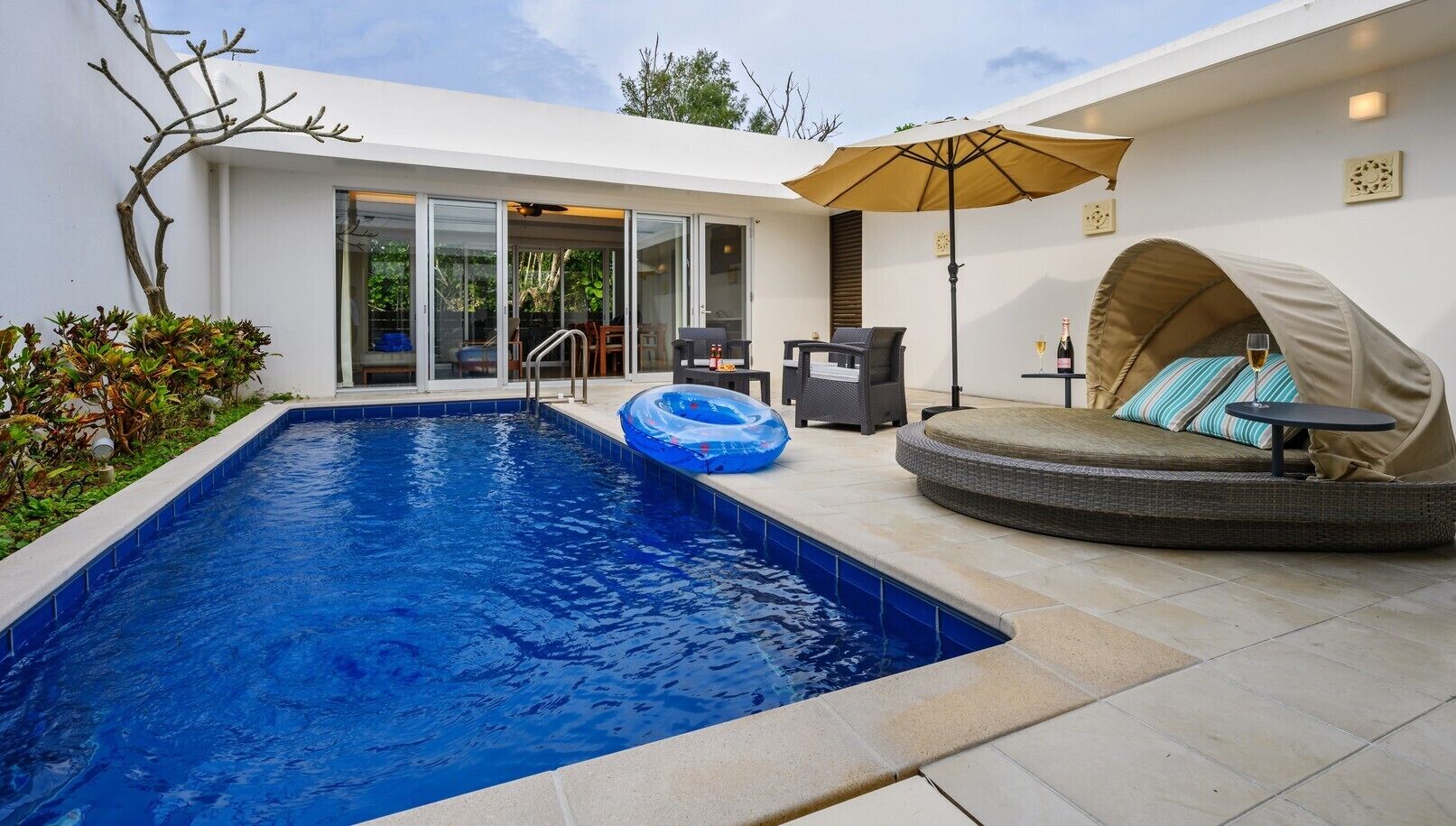 A swimming pool with a lounge area, including a covered seating area and a table with drinks.