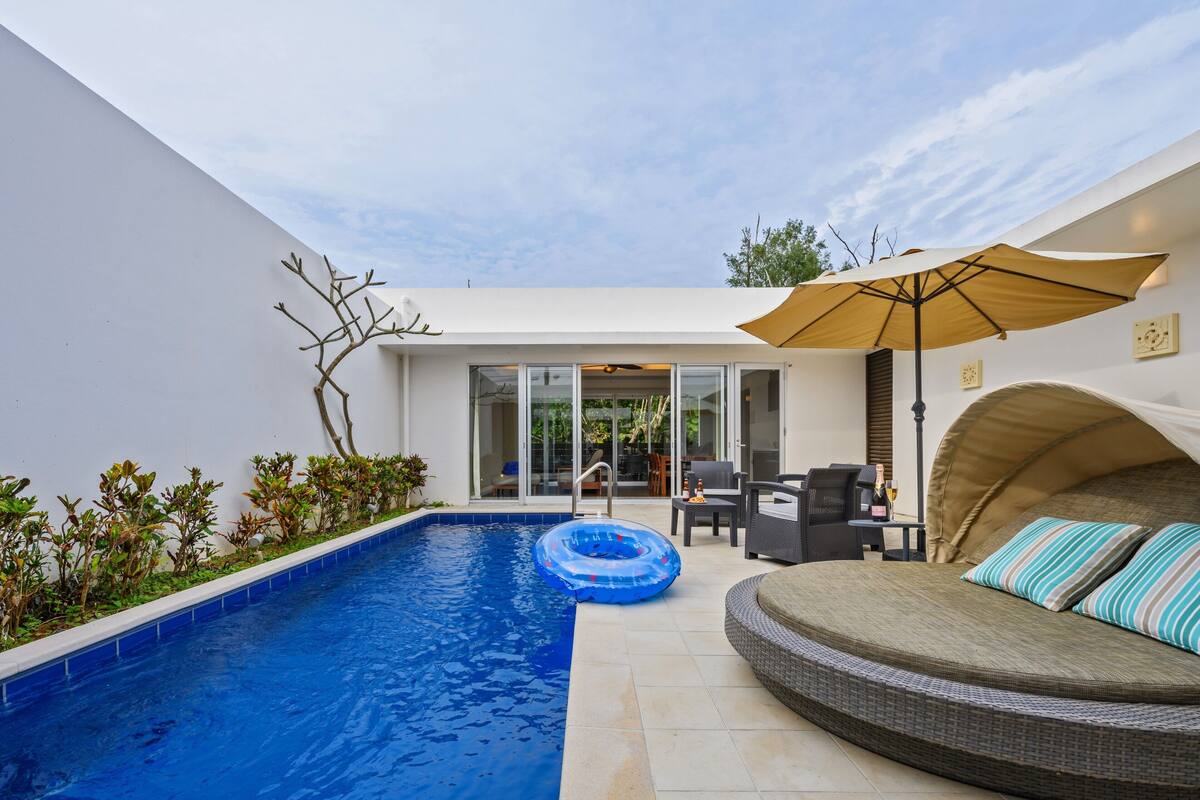 A modern outdoor pool area with a blue inflatable float, a wicker sofa with striped cushions, and a large umbrella providing shade.