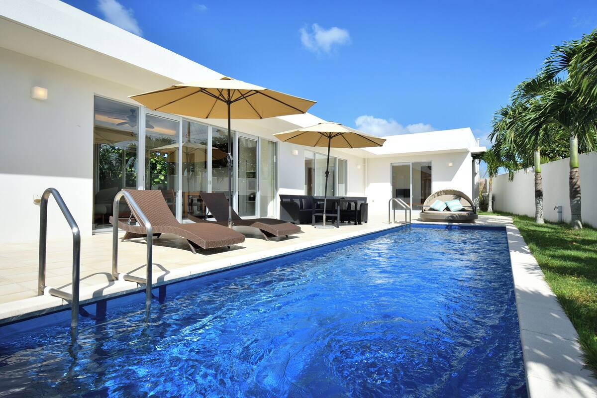 A modern swimming pool with lounge chairs and umbrellas, surrounded by a white building with large windows.