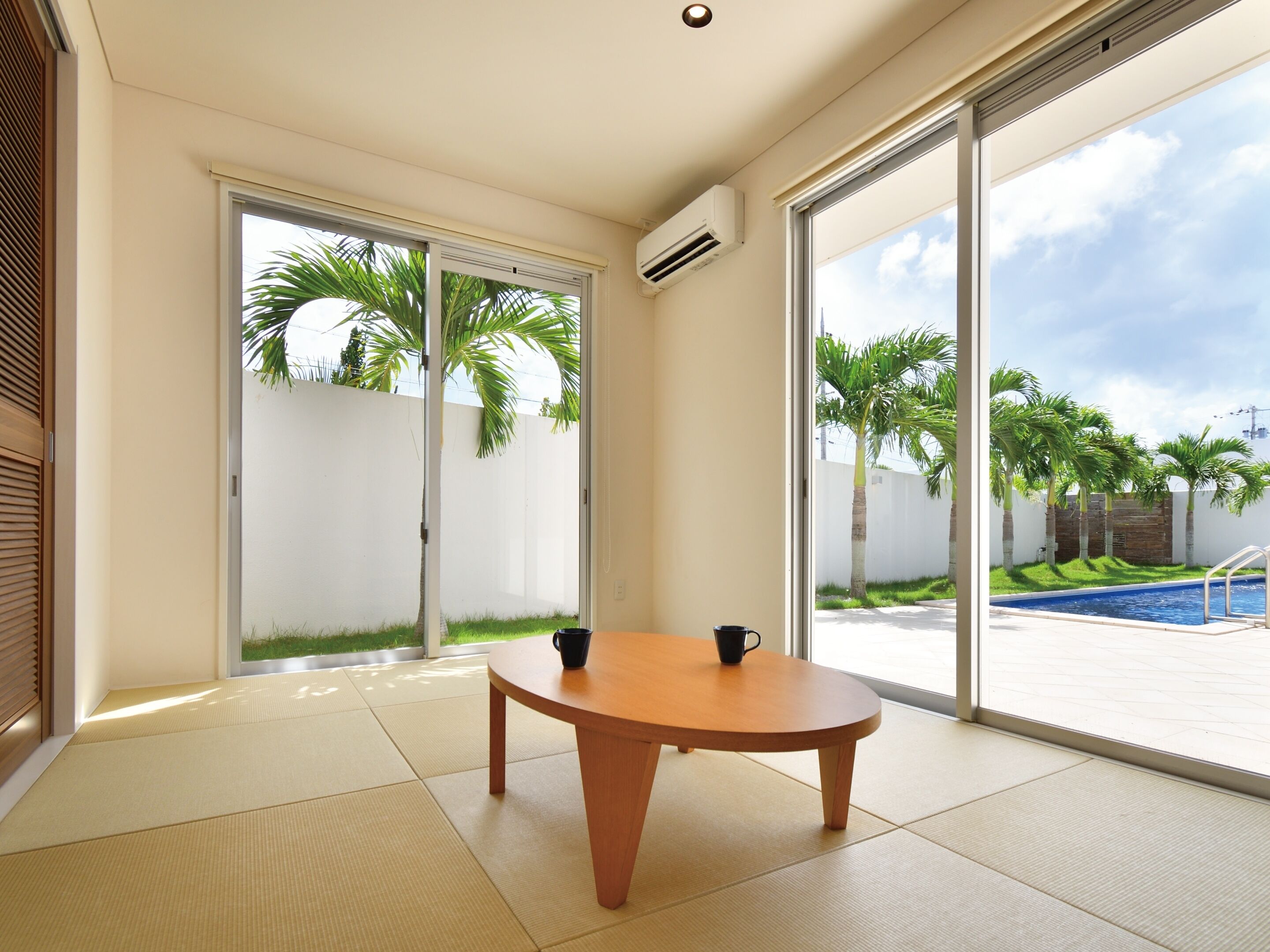 Una habitación con suelo de estera tatami, una mesa de madera con dos tazas negras, y grandes puertas correderas de cristal que dan a una zona de piscina con palmeras.