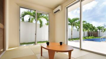 Una habitación con suelo de estera tatami, una mesa de madera con dos tazas negras, y grandes puertas correderas de cristal que dan a una zona de piscina con palmeras.