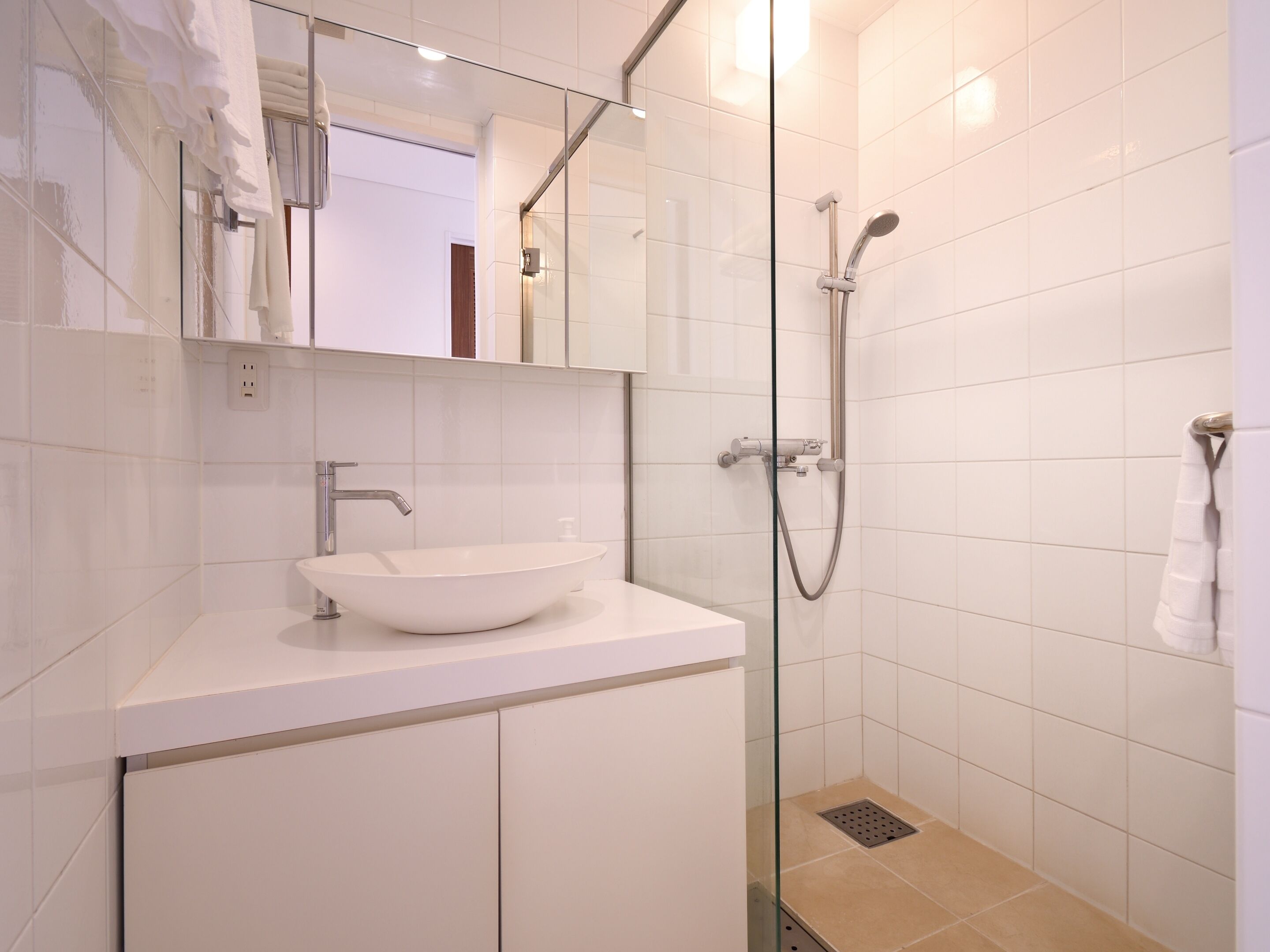 Un baño moderno con un lavamanos blanco, mampara de ducha de vidrio y pared cubierta de azulejos blancos.