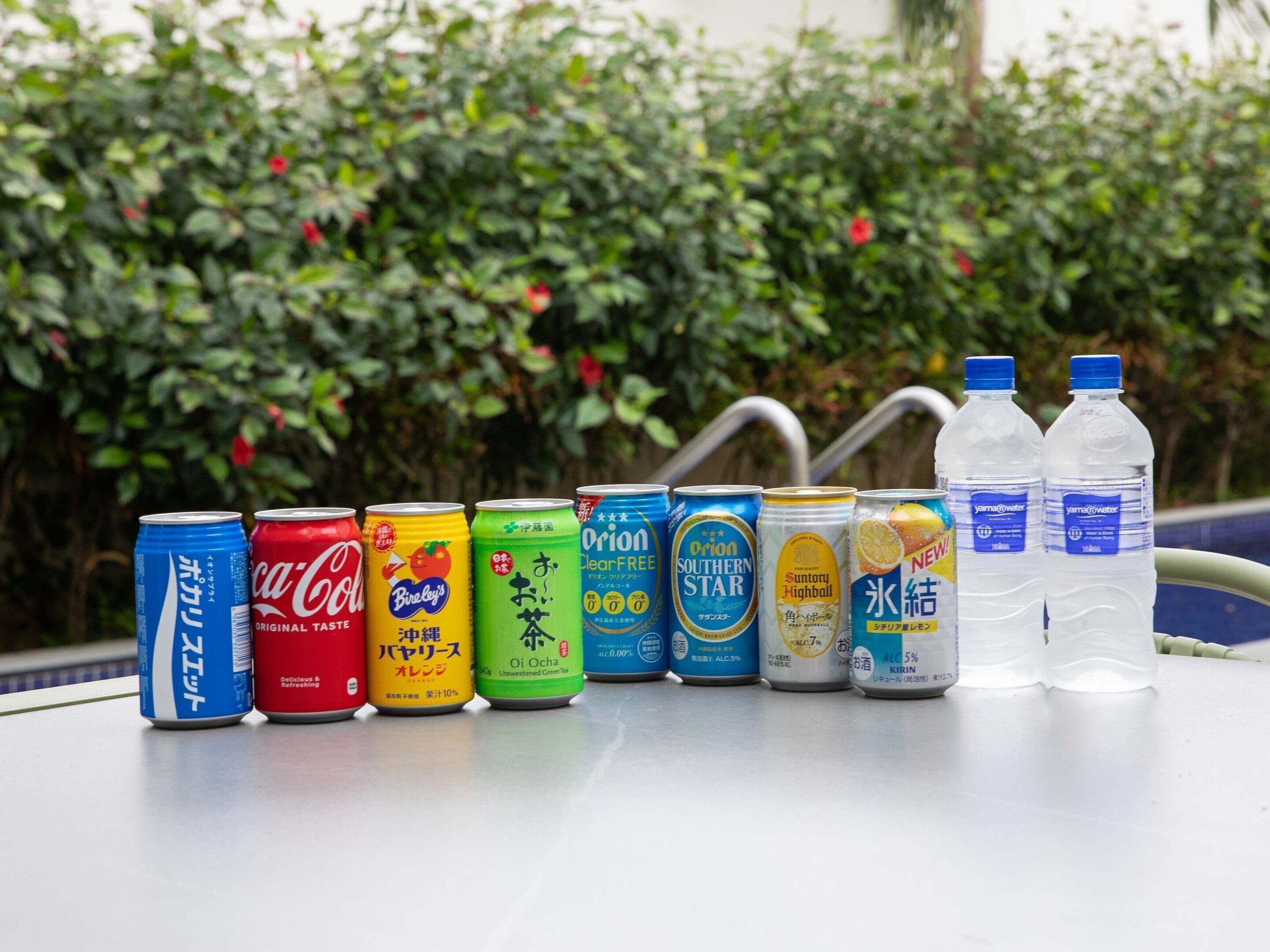 A variety of beverage cans and bottles arranged on a table outdoors with a hedge in the background.