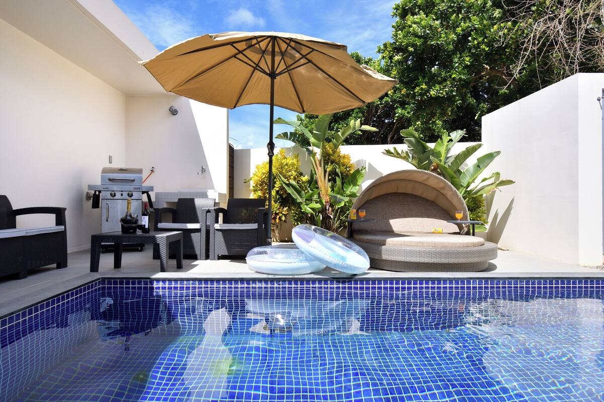 A swimming pool with blue mosaic tiles, outdoor furniture, a barbecue grill, and a large umbrella.