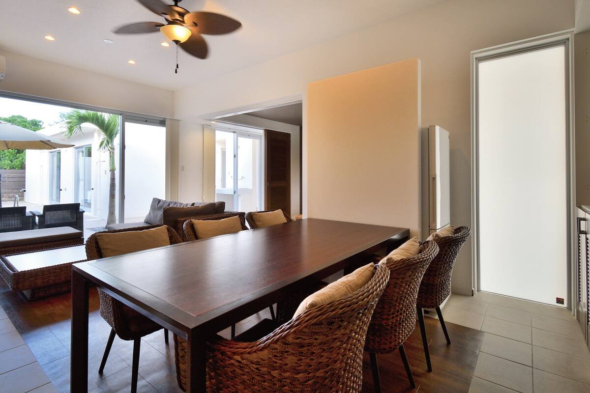 A spacious dining area with a long table and wicker chairs, a ceiling fan, and a view of the outdoors through large windows.