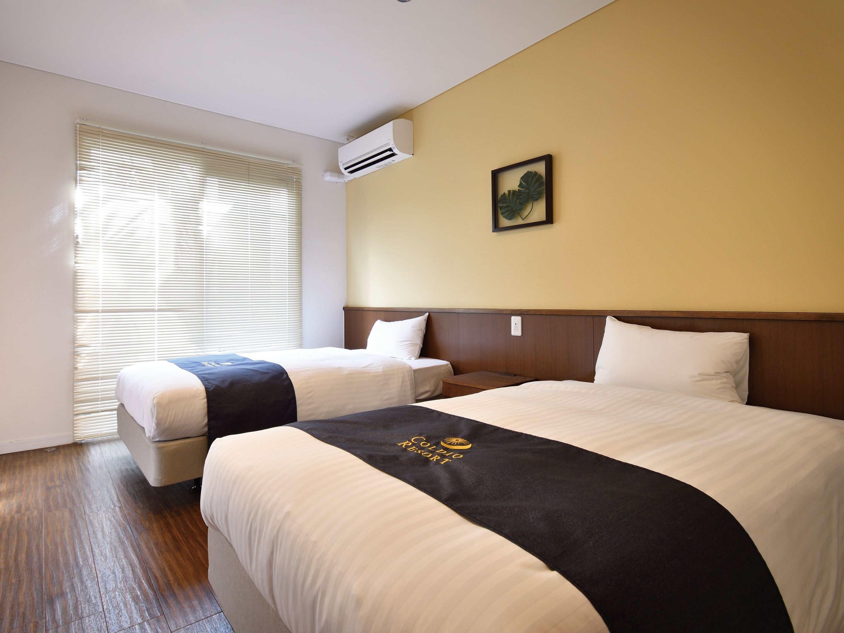 A hotel room with two beds, a large window with blinds, an air conditioning unit, and a framed plant on the wall.