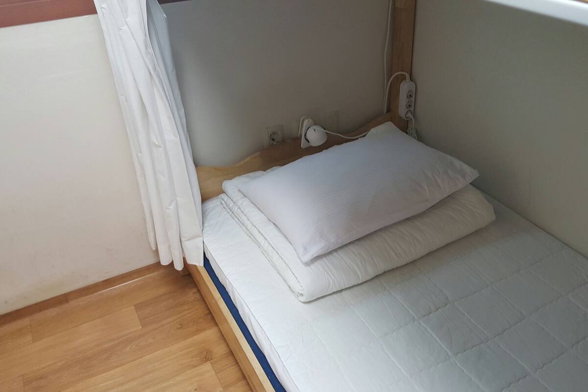A small, neatly made bed with a white mattress and pillows, a wooden headboard, and a power outlet on the wall.