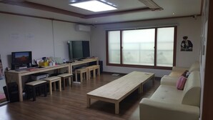 A living room with a wooden floor, a beige sofa, a wooden coffee table, a TV, and a large window with blinds. - Happy Tree Guesthouse - Hostel (Seoul)