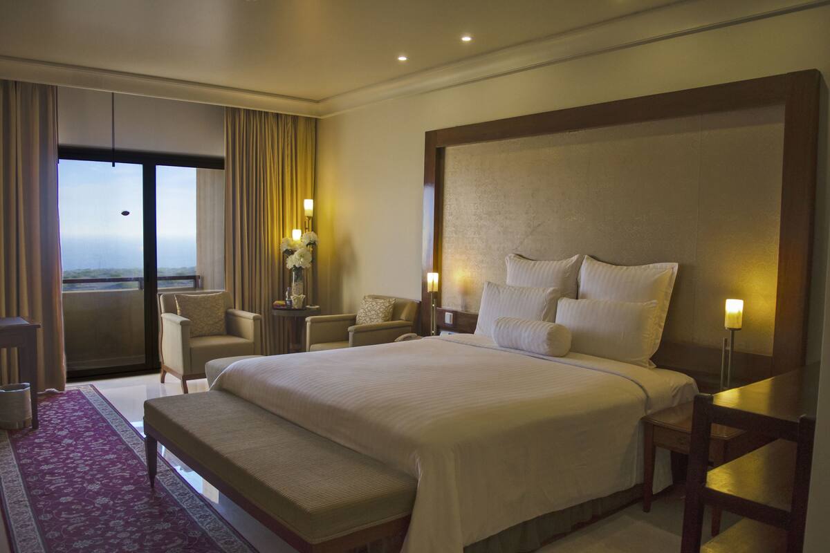 A hotel room with a large bed, a sitting area with chairs, a small table with a vase of flowers, and a view through a large window.