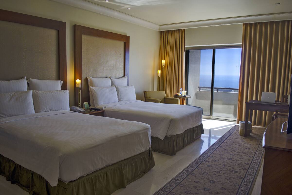 A hotel room with two beds, a desk, a chair, a large window with curtains, and a view of the sea.