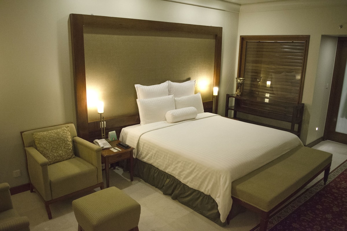 A hotel room with a large bed, bedside tables, a chair, a bench, and a window with blinds.