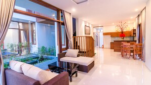 A modern living room with a sofa, a dining area with wooden chairs, a kitchen with a wooden countertop, and a view of a garden through large windows. - Green World Hoi An Villa (Da Nang)