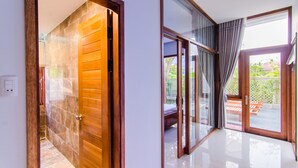 A modern interior with a wooden door leading to a bathroom, a glass door to an outdoor area, and a tiled floor. - Green World Hoi An Villa (Da Nang)