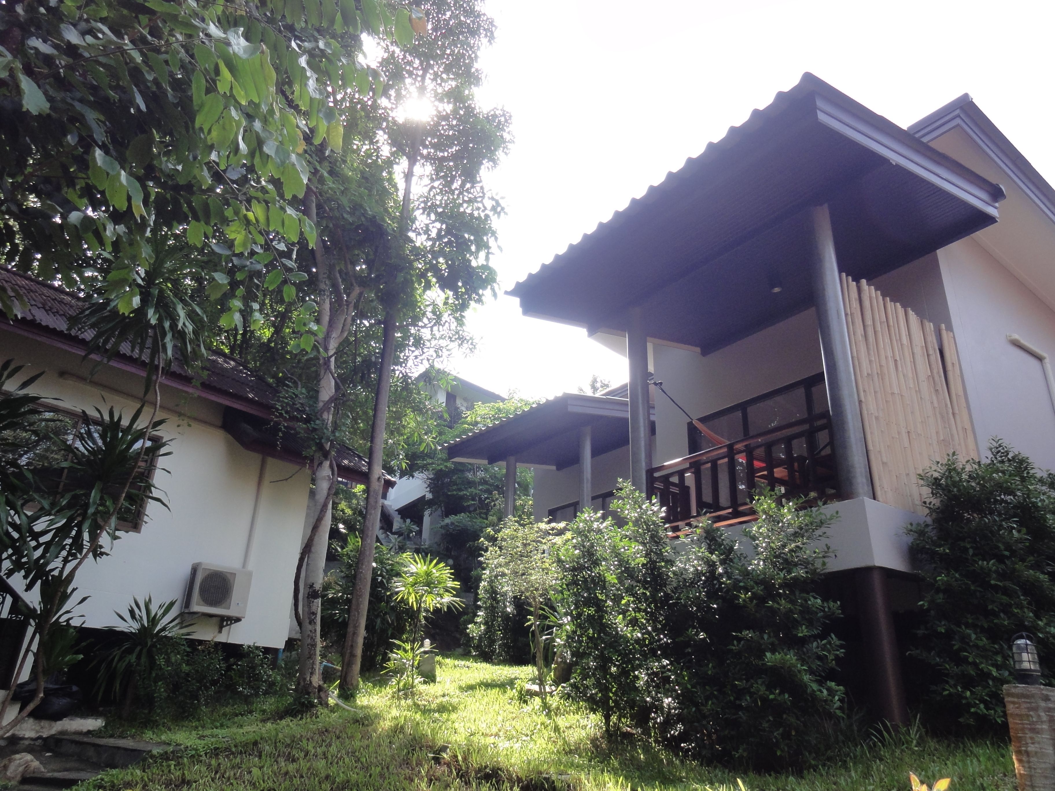 A two-story building with a covered balcony, surrounded by lush greenery and a small garden area.