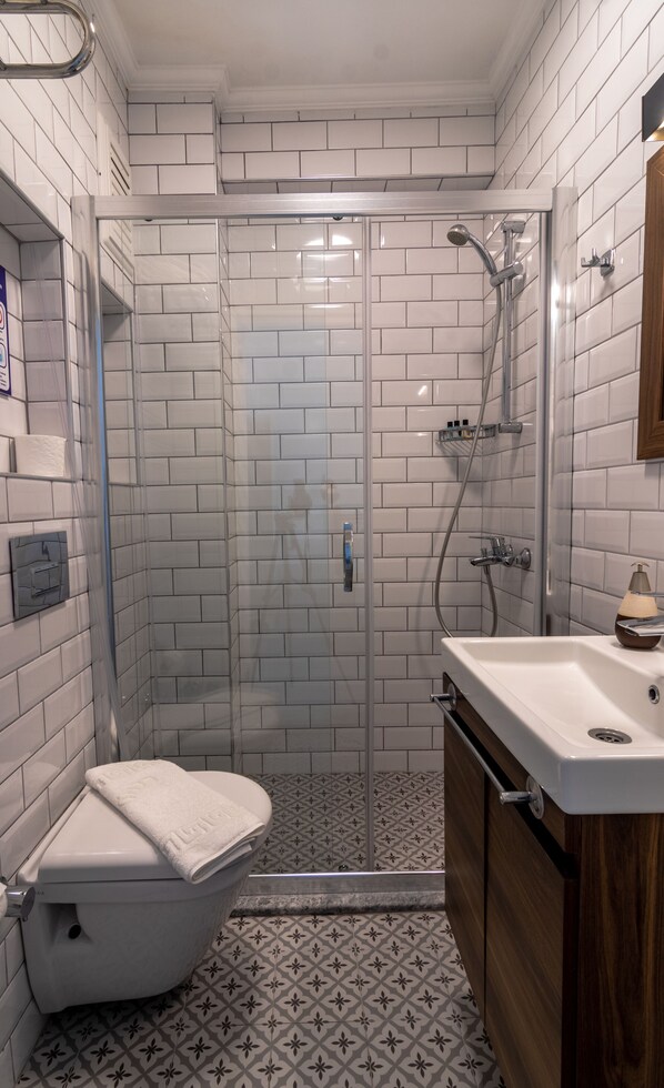 Un baño con azulejos blancos tipo metro, una ducha con puerta de vidrio, un inodoro y un lavamanos con gabinete de madera.