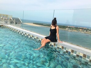 En kvinna sitter vid poolkanten med utsikt över havet och en byggnad i bakgrunden.