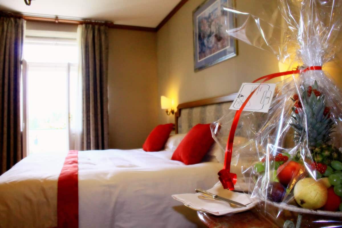 Ein Hotelzimmer mit einem ordentlich bezogenen Bett, einem Obstkorb auf dem Nachttisch und einem Fenster mit Vorhängen.