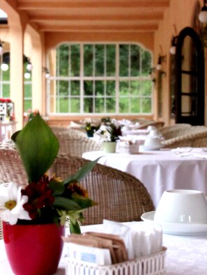 A dining area with wicker furniture, white tablecloths, and a large window with a view of greenery. - Miramonti Majestic Grand Hotel (Cortina d'Ampezzo)