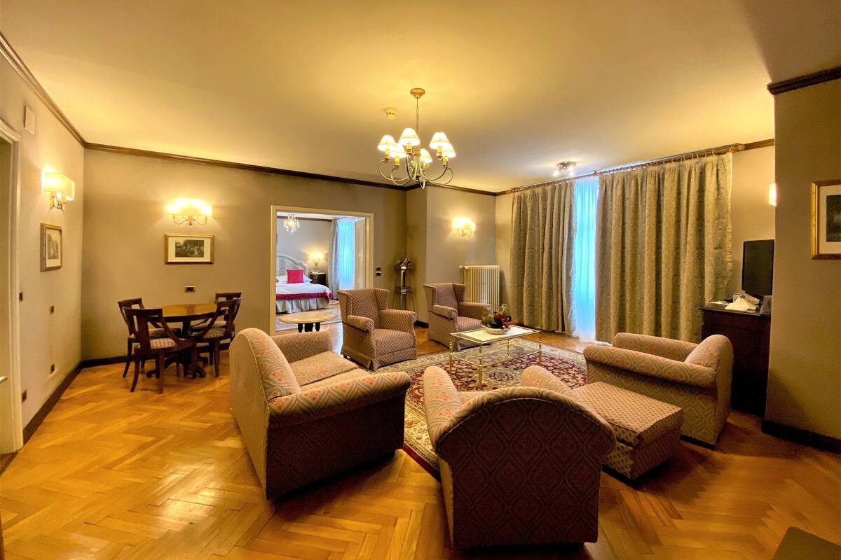Ein Wohnzimmer mit Holzboden, einer Kristallleuchter, einem Esstisch mit Stühlen und Blick auf ein Schlafzimmer durch eine geöffnete Tür.