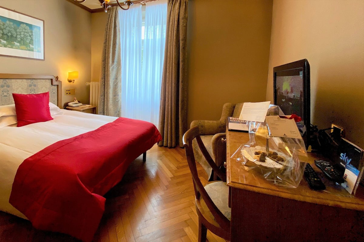 Ein Hotelzimmer mit einem Bett, einem Schreibtisch mit Computer und Fernbedienungen, einem Sessel und einem Fenster mit Vorhängen.