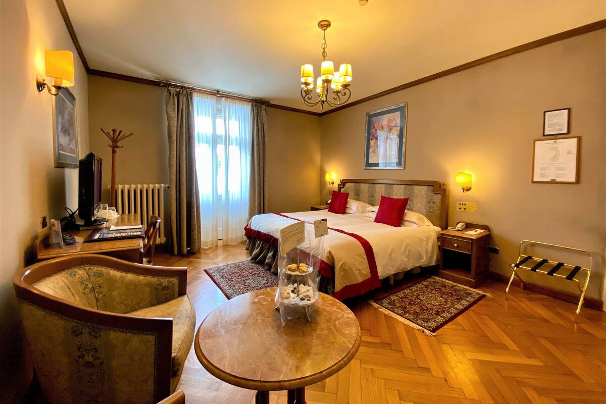 Ein Hotelzimmer mit Bett, Schreibtisch, Stuhl, Tisch, Heizkörper, einer Chandelierleuchtre und einem Wandbild.