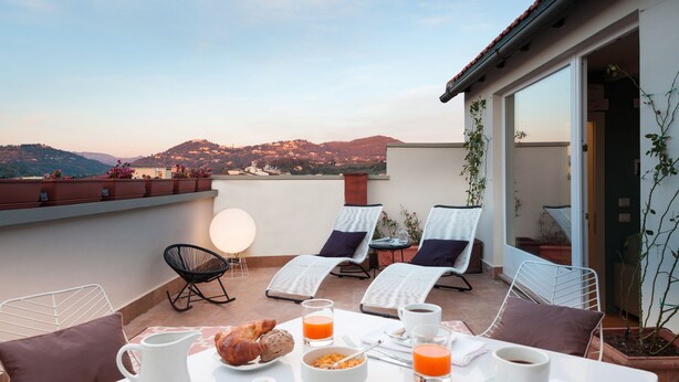 Una terraza con una mesa dispuesta para el desayuno, que incluye croissants, zumo de naranja y café, con vistas a un paisaje montañoso.