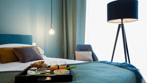 Habitación de hotel con una cama bien tendida, una mesita de noche con artículos de desayuno, una lámpara de pie y vistas al exterior.