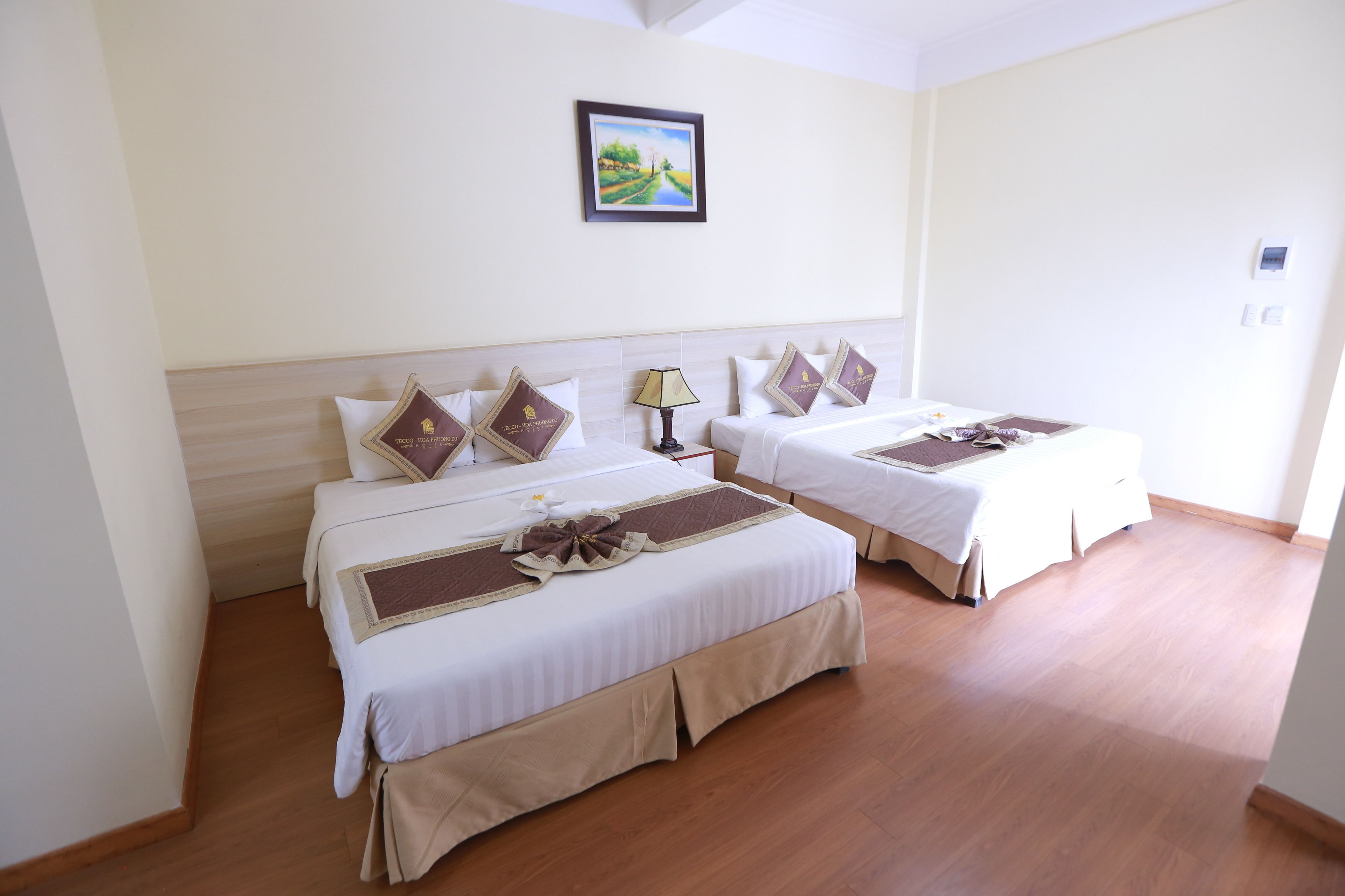 A hotel room with two beds, each with a brown decorative pillow, a bedside lamp, and a framed picture on the wall.