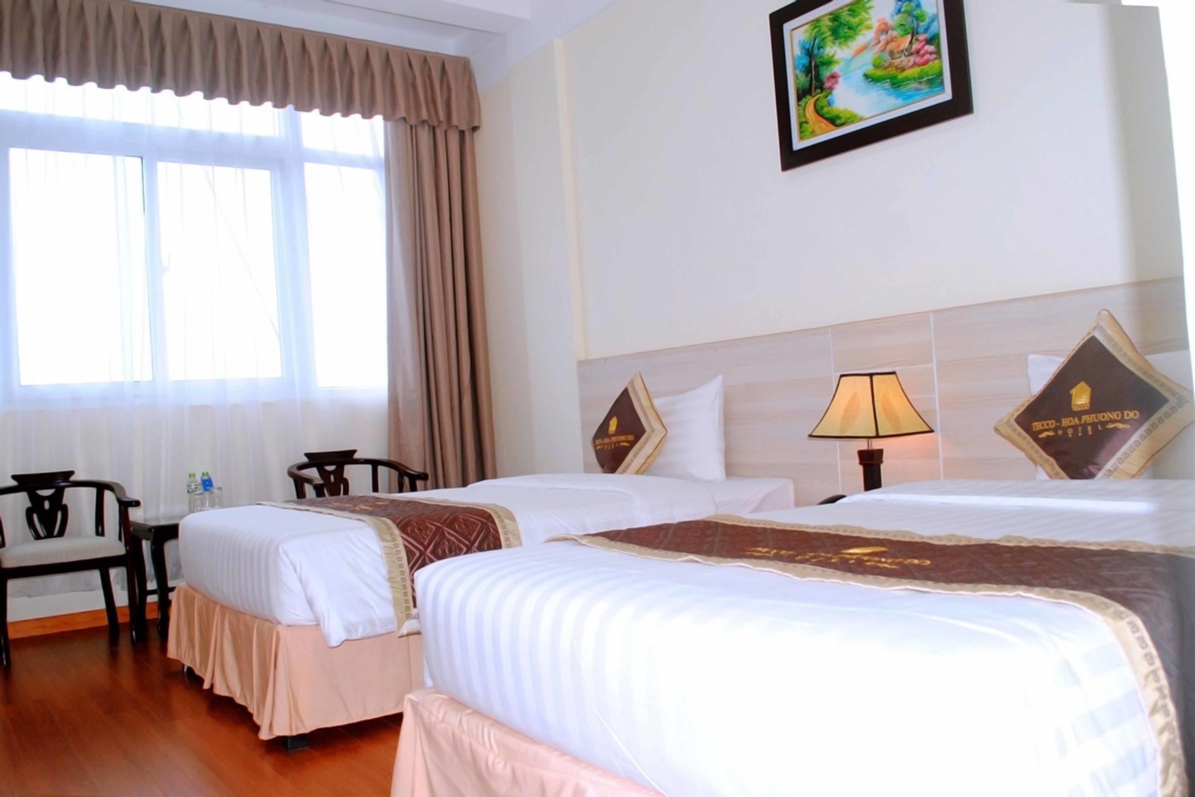 A hotel room with two beds, a desk, chairs, a lamp, and a painting on the wall.