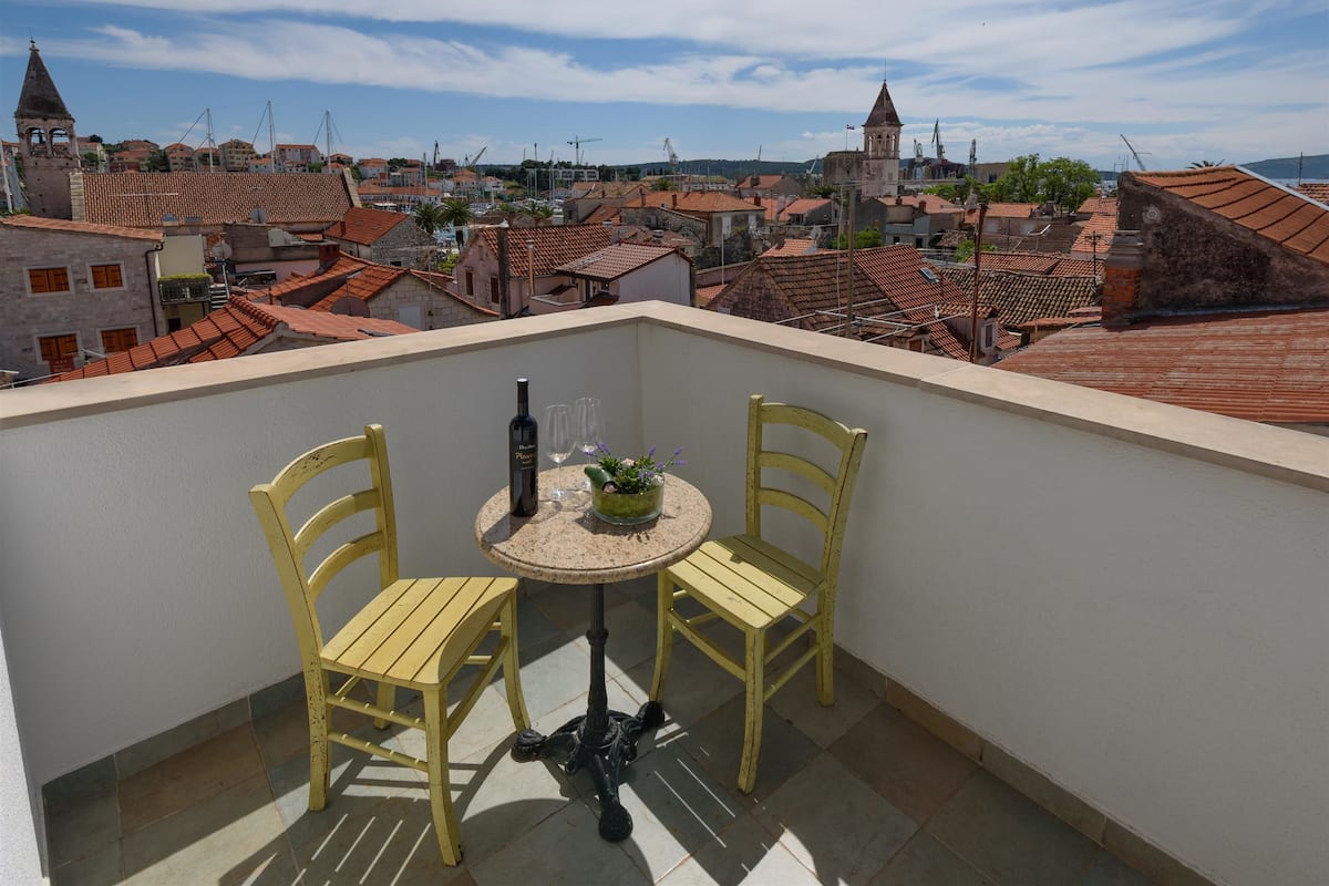 A balcony with two yellow chairs and a small round table, overlooking a cityscape with red-roofed buildings and a church tower.