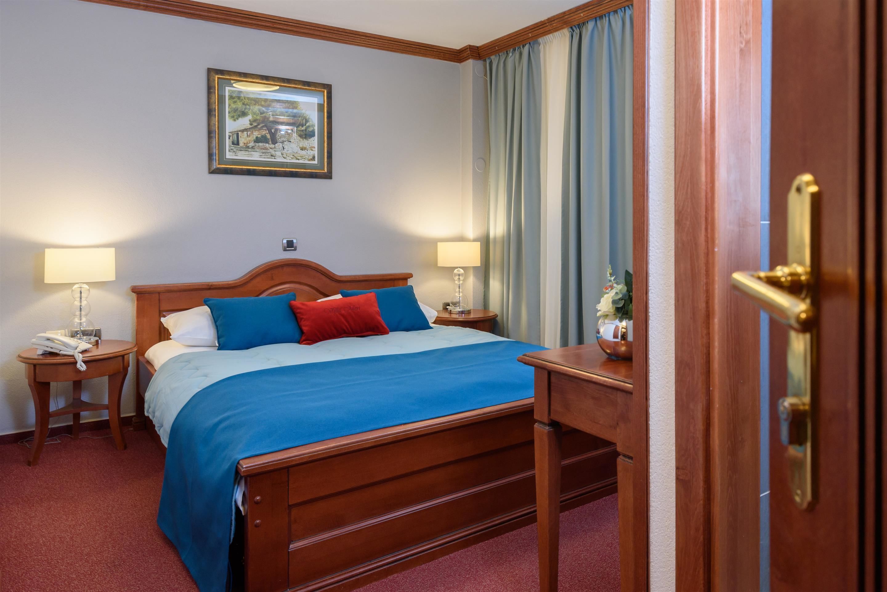 A hotel room with a wooden bed, two bedside tables with lamps, a nightstand with a vase of flowers, and a framed picture on the wall.