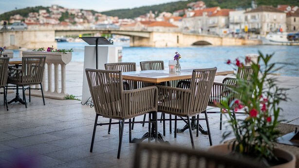 An outdoor dining area with wicker chairs and a table, overlooking a river with a bridge and buildings in the background. - Villa Meri (Trogir)