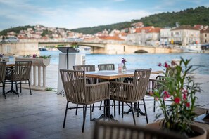 Un espace extérieur aménagé pour les repas, avec des chaises et une table en osier, donnant sur une rivière, un pont et des bâtiments en arrière-plan.