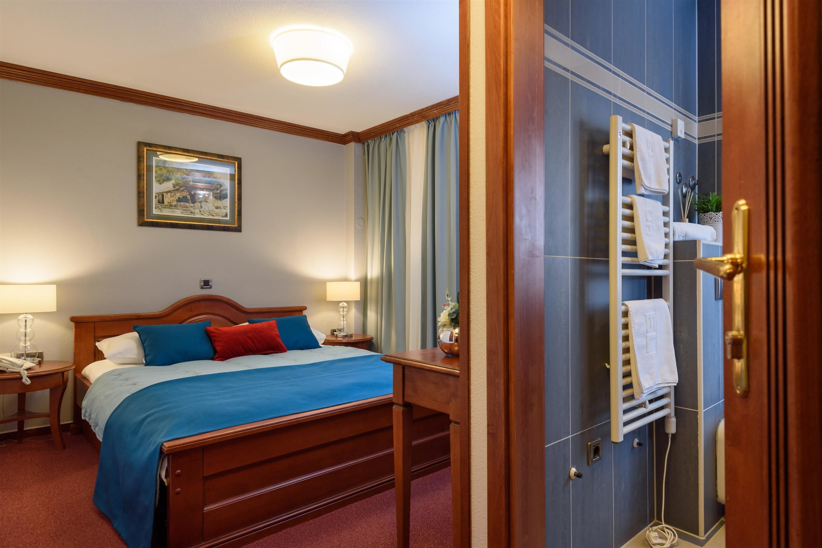 A hotel room with a wooden bed, a nightstand with a lamp, a framed picture on the wall, and a bathroom with a towel rack.