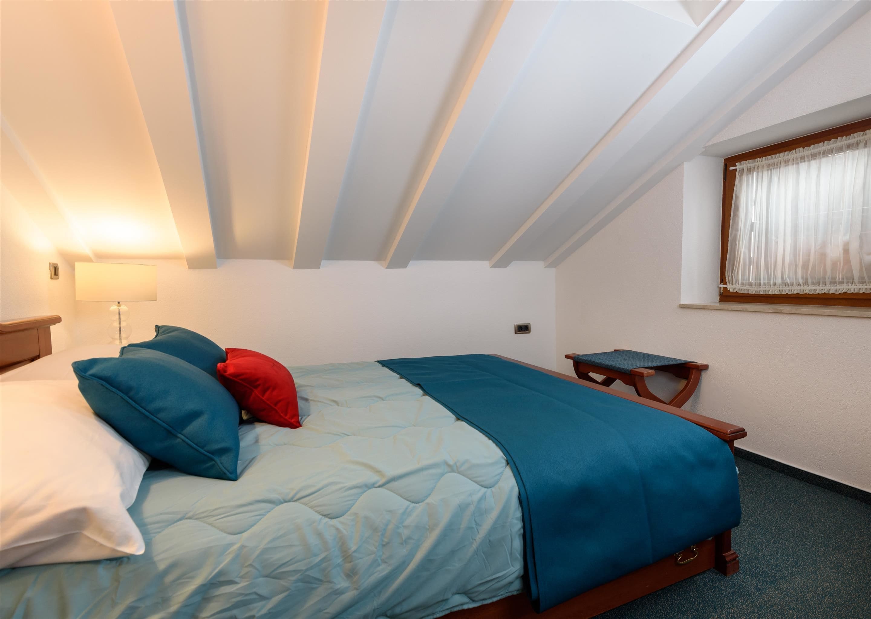 Ein Einzelbett mit blauer Bettdecke und Kissen, ein Nachttisch mit Lampe sowie ein Fenster mit Vorhängen.