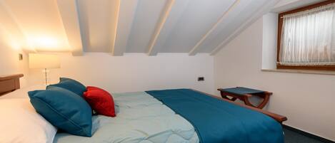 Un lit simple avec une couvre-lit et des oreillers bleus, une table de chevet avec une lampe, et une fenêtre avec des rideaux.